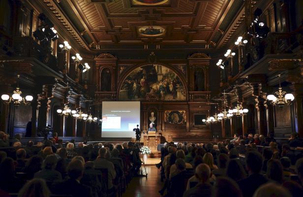 Aufnahme der Alten Aula der Uni Heidelberg von der Eingangstür des Saals aus. Das Foto zeigt über die Köpfe des Publikums hinweg den holzvertäfelten Saal. Die Frontseite weist ein halbrundes Gemälde auf, vor dem ein Rednerpult steht. Neben dem Pult ist eine Leinwand heruntergelassen.