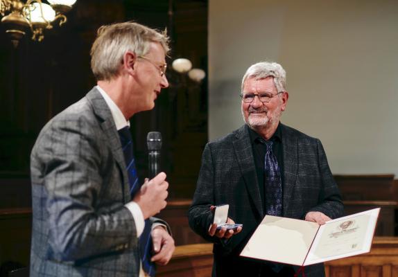 Halbnahaufnahme von Robert Schlögl und dem Akademiepräsidenten 2023 Hans-Georg Kräusslich. Herr Kräusslich ist von der Seite aufgenommen, hält ein Mikrofon in der Hand und wendet sich Herrn Schlögl zu, der eine aufgeschlagene Urkunde in der Hand hält und eine Schatulle mit der Akademiemedaille. Herr Schlögl ist frontal aufgenommen und lächelt Herrn Kräusslich an.