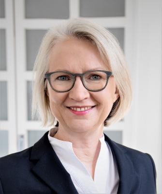 Portrait von Monika Schnitzer in Nahaufnahme, sie trägt eine weiße Bluse, darüber einen dunkelblauen Blazer, ihr weiß-blondes Haar trägt sie als Bob, dazu eine dunkelgerahmte Hornbrille