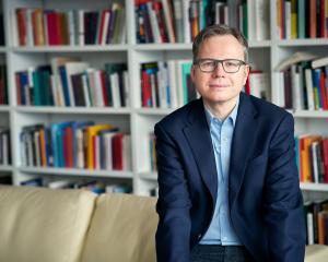 Portrait von Andreas Reckwitz hüftaufwärts. Er sitzt vor einem Bücherregal auf der Lehne eines hellen Sofa. Er trägt ein dunkelblaues Jackett, darunter ein hellblaues Hemd. Dazu eine dunkel gerahmte, eckige Brille.