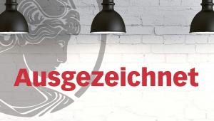 Das Bild zeigt eine weiß gekachelte Wand vor der drei schwarze Lampenschirme hängen. Sie beleuchten das in roter Schrift abgebildete Wort "Ausgezeichnet". In der linken Bildhälfte ist das Logo der Akademie, die Athene im Seitenprofil, zu erkennen, die in grau transparent an die Wand gemalt ist.