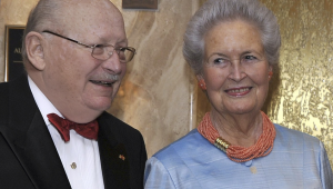 Portrait von Sigrid und Viktor Dulger in halbnaher Aufnahme. Links im Bild steht Herr Dulger, rechts von ihm seine Frau. Herr Dulger trägt ein weißes Hemd, eine rote Fliege und darüber ein schwarzes Jackett. Dazu eine eckige, metallgerahmte Brille. Sein Kopf ist nahezu kahl und er lächelt mit nach rechts gerichtetem Gesicht einer nicht zu sehenden Person zu. Frau Dulger trägt einen hellblauen Blazer, dazu eine korallfarbene Perlenkette und passende Ohrringe in Blütenform. Ihr Blick ist mach links gerichtet.